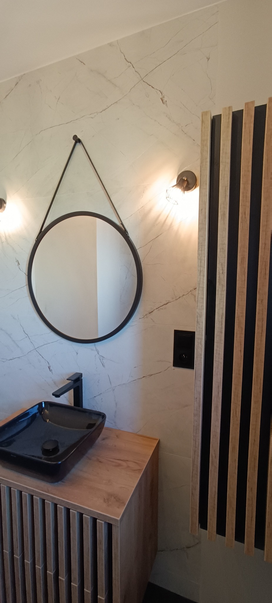 Wykończona łazienka z czarną umywalką nablatową, okrągłym lustrem na tle marmurowych płytek i drewnianymi lamelami. Nowoczesny design z oświetleniem punktowym.