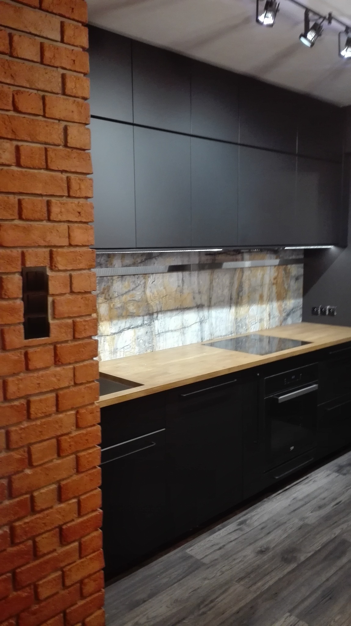 Nowoczesna kuchnia z czarnymi szafkami, drewnianym blatem i ceglaną ścianą z wnęką. Oświetlenie punktowe podkreśla marmurowy backsplash.