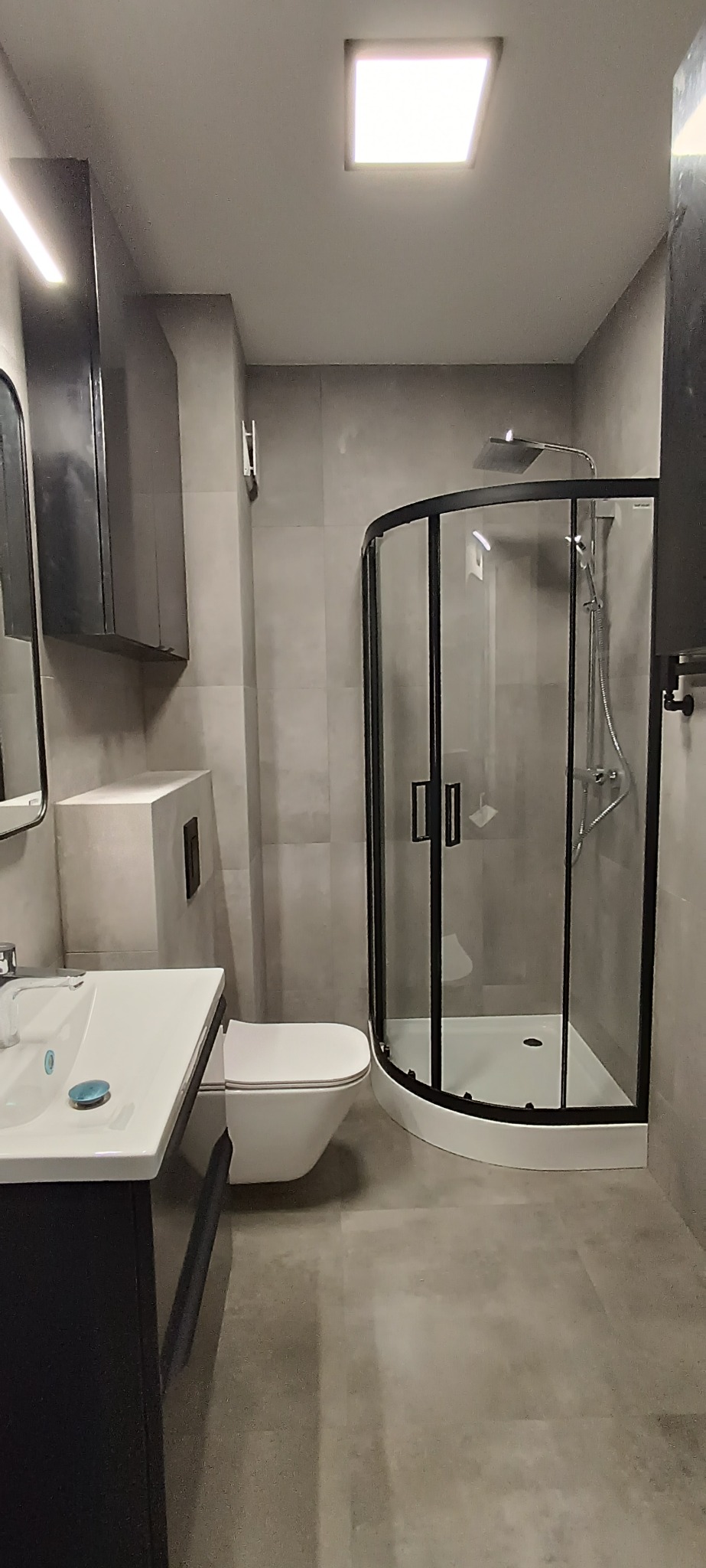 Nowoczesna łazienka w szarościach z kabiną prysznicową z czarnymi ramami, umywalką z szafką i podwieszaną toaletą. Minimalistyczny design, kwadratowe płytki.