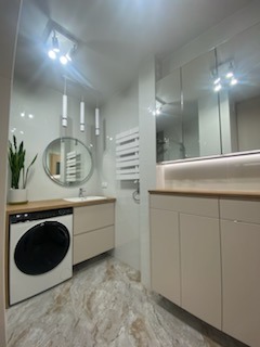 Jasna łazienka z marmurową podłogą, okrągłym lustrem i pralką obok szafki umywalkowej. Minimalistyczne oświetlenie punktowe i grzejnik drabinkowy.