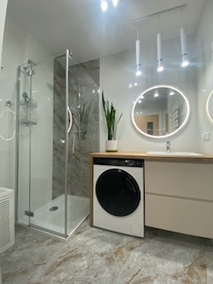 Nowoczesna łazienka z kabiną prysznicową, pralką pod blatem i okrągłym lustrem z podświetleniem LED. Uwagę zwraca marmurowa podłoga i minimalistyczne oświetlenie.