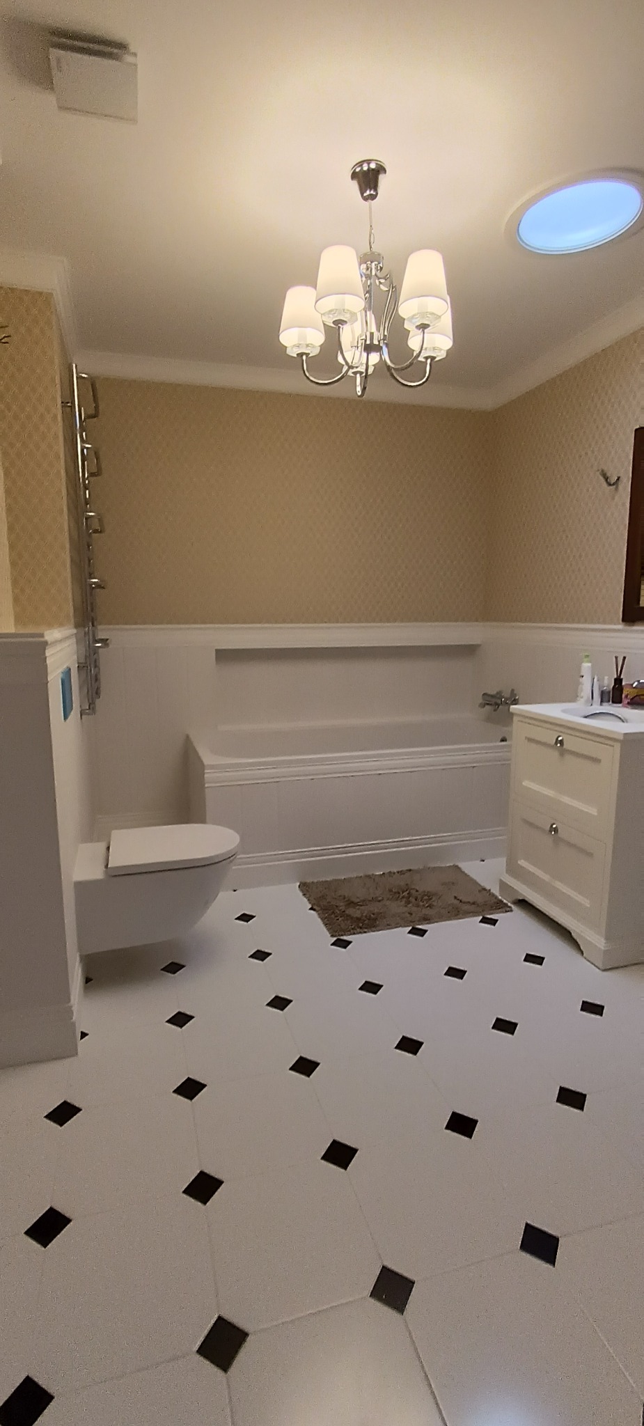 Elegancka łazienka w jasnych barwach z białą armaturą, czarnymi akcentami na podłodze i ozdobnym żyrandolem. Widoczna wanna, toaleta i szafka z umywalką.