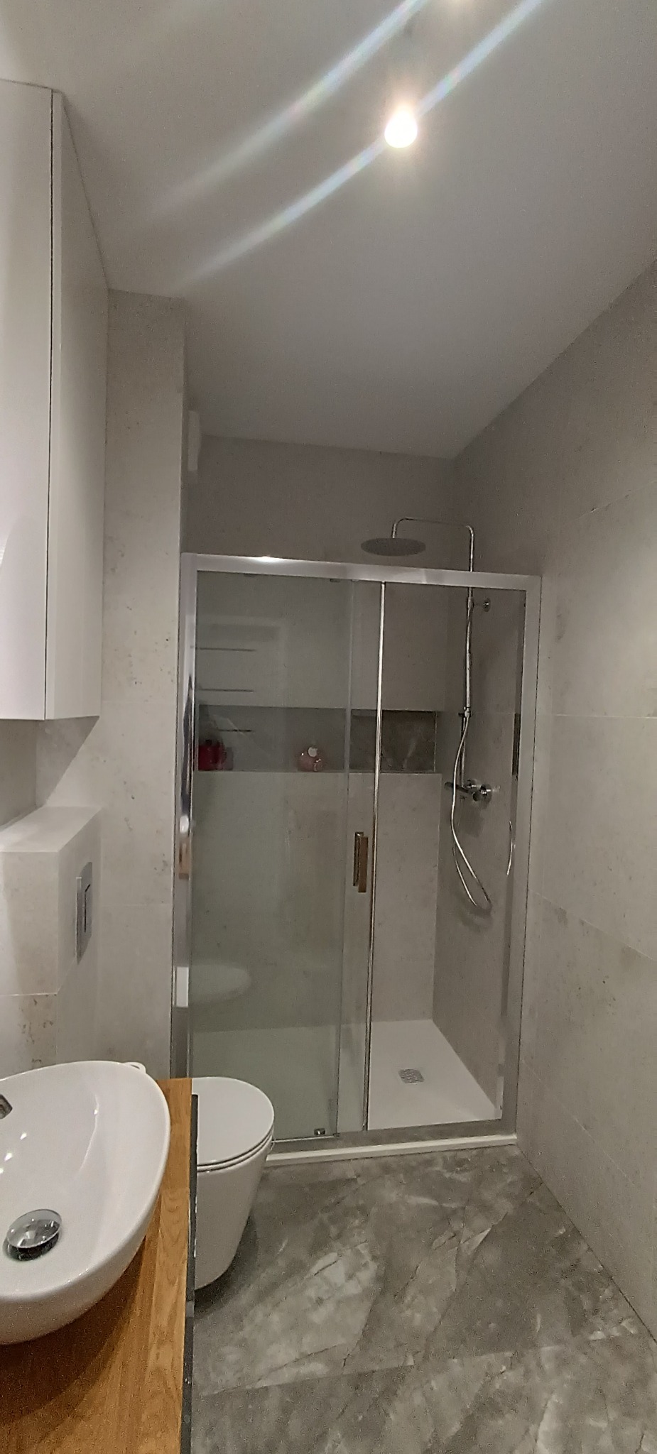 Nowoczesna łazienka z kabiną prysznicową typu walk-in, umywalką na drewnianym blacie i szarą marmurową podłogą. Minimalistyczny design, jasne płytki na ścianach.