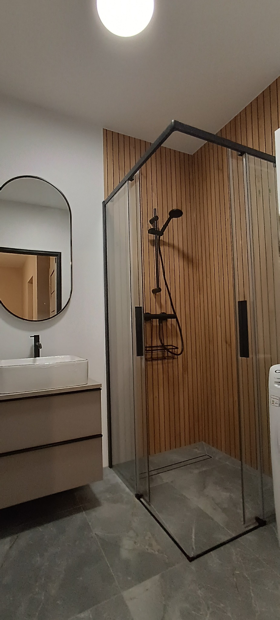 Nowoczesna łazienka z kabiną prysznicową typu walk-in, umywalką nablatową i lustrem w czarnej ramie. Ściana wykończona drewnianymi panelami. Szare płytki podłogowe.