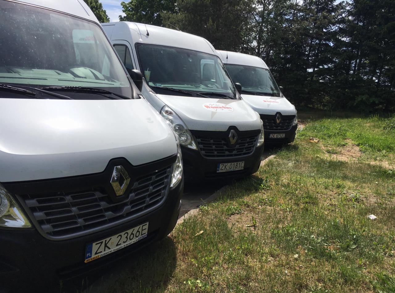 Trzy białe busy Renault Master zaparkowane obok siebie na trawiastym terenie, widoczne tablice rejestracyjne i logo firmy na środkowym busie.