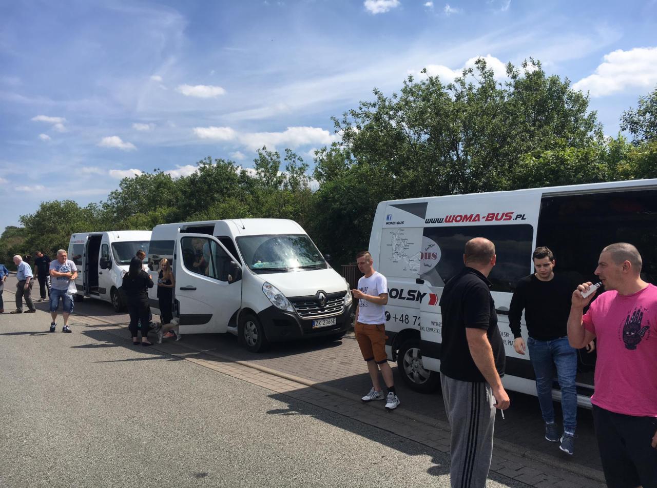 Białe busy firmy Woma-Bus zaparkowane na parkingu, pasażerowie wsiadają i wysiadają, widoczne logo firmy na busie.