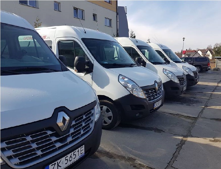 Szereg białych busów Renault Master zaparkowanych na betonowym placu przed budynkiem, logo firmy widoczne na boku pojazdu.