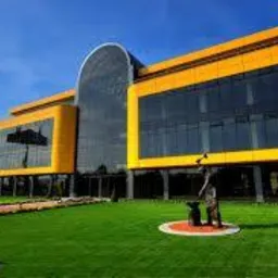 Nowoczesny budynek biurowy z żółtymi akcentami i szklaną fasadą, otoczony zielonym trawnikiem. Na pierwszym planie rzeźba kowala przy kowadle.