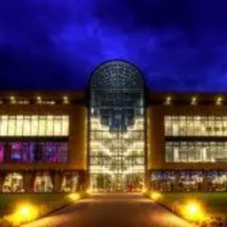 Oświetlony budynek biurowy z przeszkloną fasadą i łukowym dachem na tle nocnego nieba. Widoczne zielone trawniki i ścieżka prowadząca do wejścia.