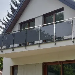 Balkon z nowoczesną balustradą ze stali nierdzewnej i szkła grafitowego, widok z dołu na tle jasnej elewacji domu z brązowymi oknami.