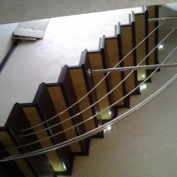 Balusmet Slawomir Janiak - Klatka schodowa z ciemnymi drewnianymi stopniami i podświetleniem LED, zabezpieczona poręczą z cienkich, zakrzywionych, metalowych prętów.