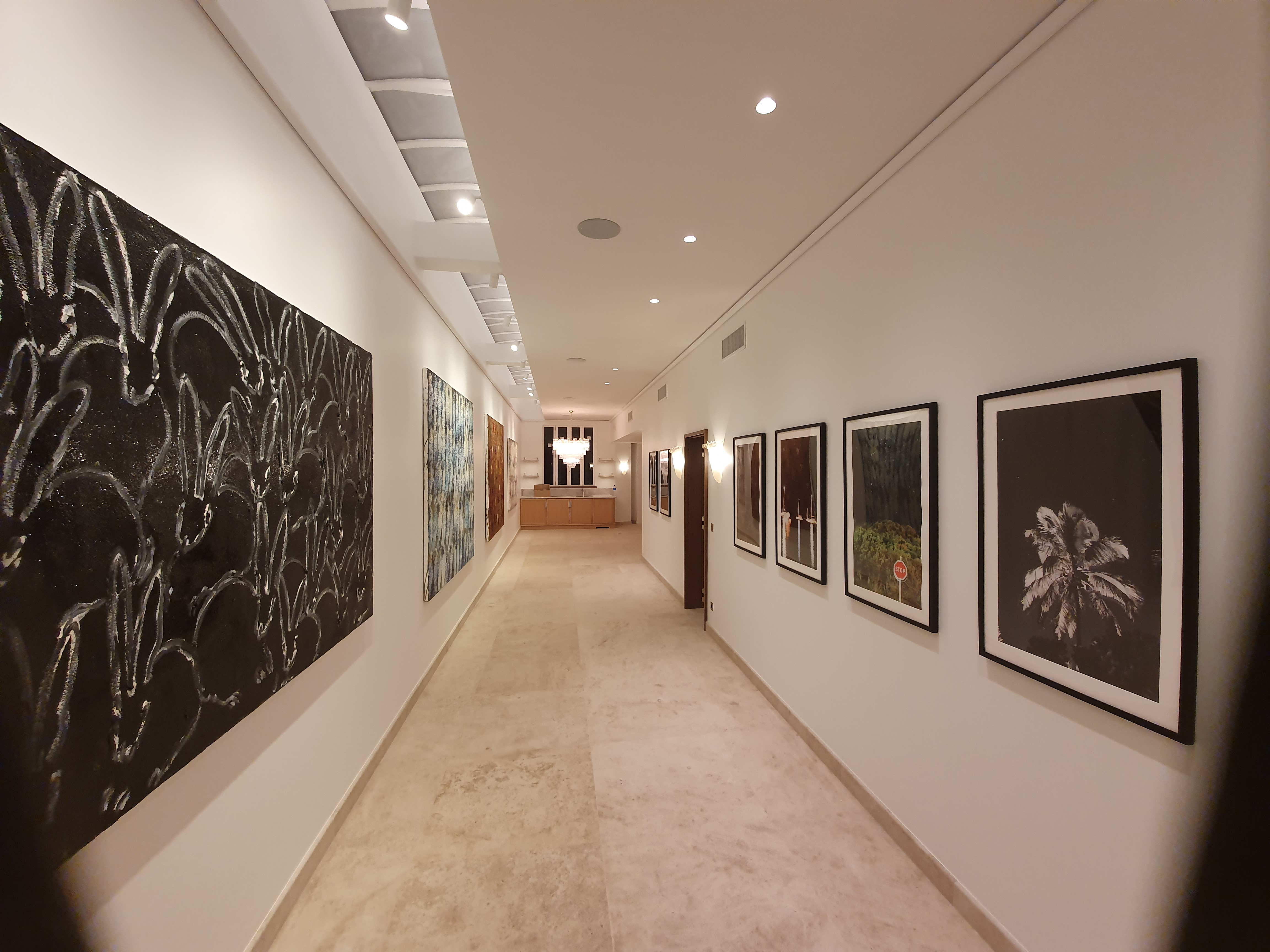 Długi korytarz w jasnych barwach z kamienną posadzką i białymi ścianami, udekorowany nowoczesnymi obrazami i oświetleniem punktowym.