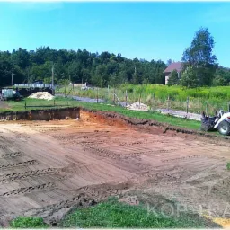 Przygotowany wykop pod fundamenty z widocznymi śladami opon i równiarką Terex na skraju, otoczony zielenią i drzewami w słoneczny dzień.