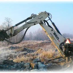 Biała koparko-ładowarka z młotem hydraulicznym kruszy betonowe elementy na zewnątrz, w tle widoczne drzewa i pole z suchą trawą, w prawym dolnym rogu logo firmy KOP-TRANS.