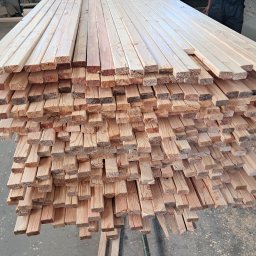 Usługi Stolarskie Patryk Torczyński - Stos świeżej, jasnej tarcicy budowlanej ułożonej warstwami, widoczny fragment osoby w tle. Drewno z widocznymi słojami, przygotowane do dalszej obróbki.