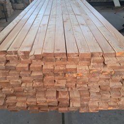 Usługi Stolarskie Patryk Torczyński - Stos tarcicy budowlanej z widocznymi słojami drewna i pomarańczowymi oznaczeniami, ułożony równo na szarym podłożu w hali produkcyjnej.