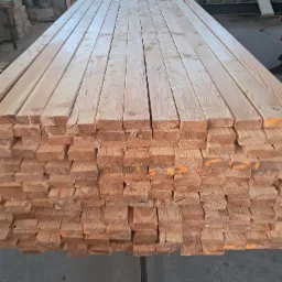 Stos tarcicy budowlanej z widocznymi słojami drewna i pomarańczowymi oznaczeniami, ułożony równo na szarym podłożu w hali produkcyjnej.