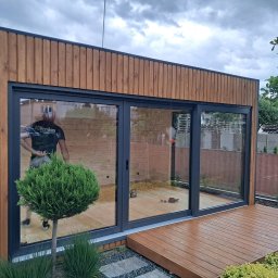 Usługi Stolarskie Patryk Torczyński - Nowoczesna, przeszklona altana z drewna, z przesuwnymi drzwiami i drewnianym tarasem. Widoczny fragment ogrodu i odbicie osoby w szybie.