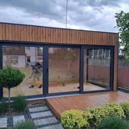 Usługi Stolarskie Patryk Torczyński - Nowoczesna altana z drewna i przeszkleniami, widoczna z zewnątrz. Taras z desek, rośliny ozdobne, ścieżka z kamieni. Prace wykończeniowe wewnątrz.