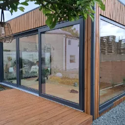 Nowoczesna altana z dużymi oknami przesuwnymi i drewnianym wykończeniem, zlokalizowana w Biskupicach, otoczona zielenią i żwirowym podłożem.