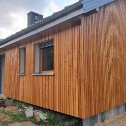 Usługi Stolarskie Patryk Torczyński - Nowoczesny dom szkieletowy z elewacją z pionowo ułożonych desek w ciepłym, miodowym odcieniu drewna. Widoczne okna i otwarte drzwi wejściowe. W tle komin i fragment rusztowania.
