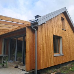 Usługi Stolarskie Patryk Torczyński - Nowoczesny dom szkieletowy z drewnianą elewacją i pergolą, widok z zewnątrz. Detale: rynna, okna, narzędzia na placu budowy. Jasne drewno, metalowy dach.