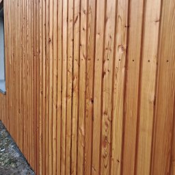 Usługi Stolarskie Patryk Torczyński - Elewacja budynku wykończona pionowymi deskami z jasnego drewna, ukazująca naturalny rysunek słojów. Widoczny fragment okna z parapetem i przygruntowy teren.