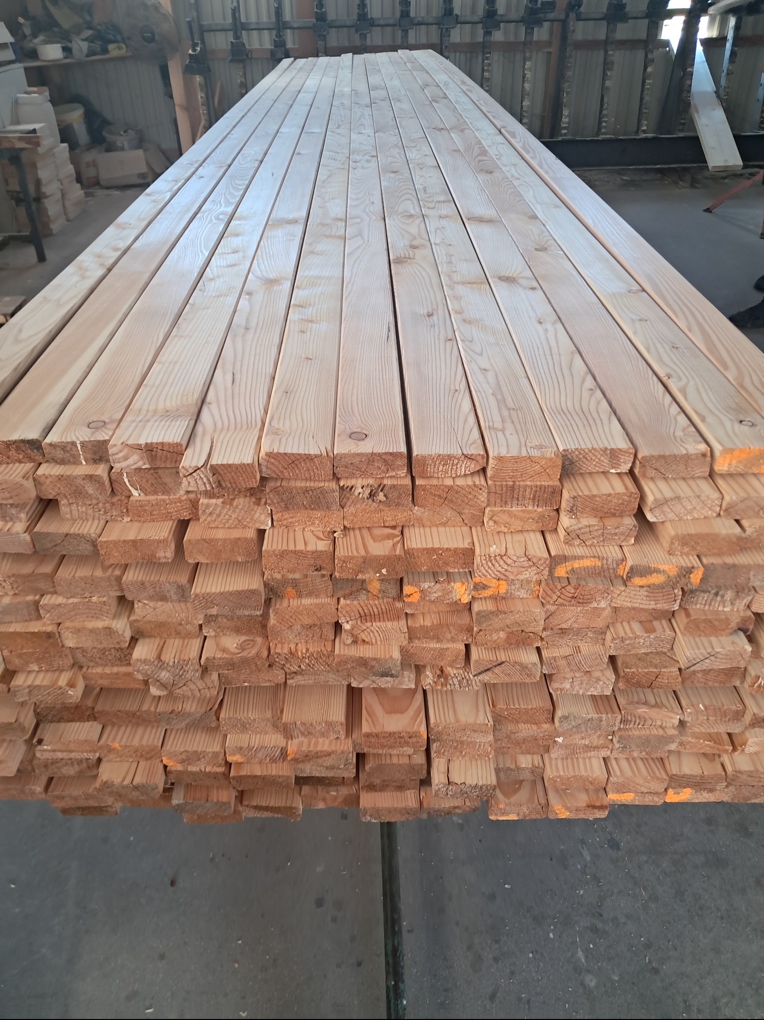 Stos tarcicy budowlanej z widocznymi słojami drewna i pomarańczowymi oznaczeniami, ułożony równo na szarym podłożu w hali produkcyjnej.