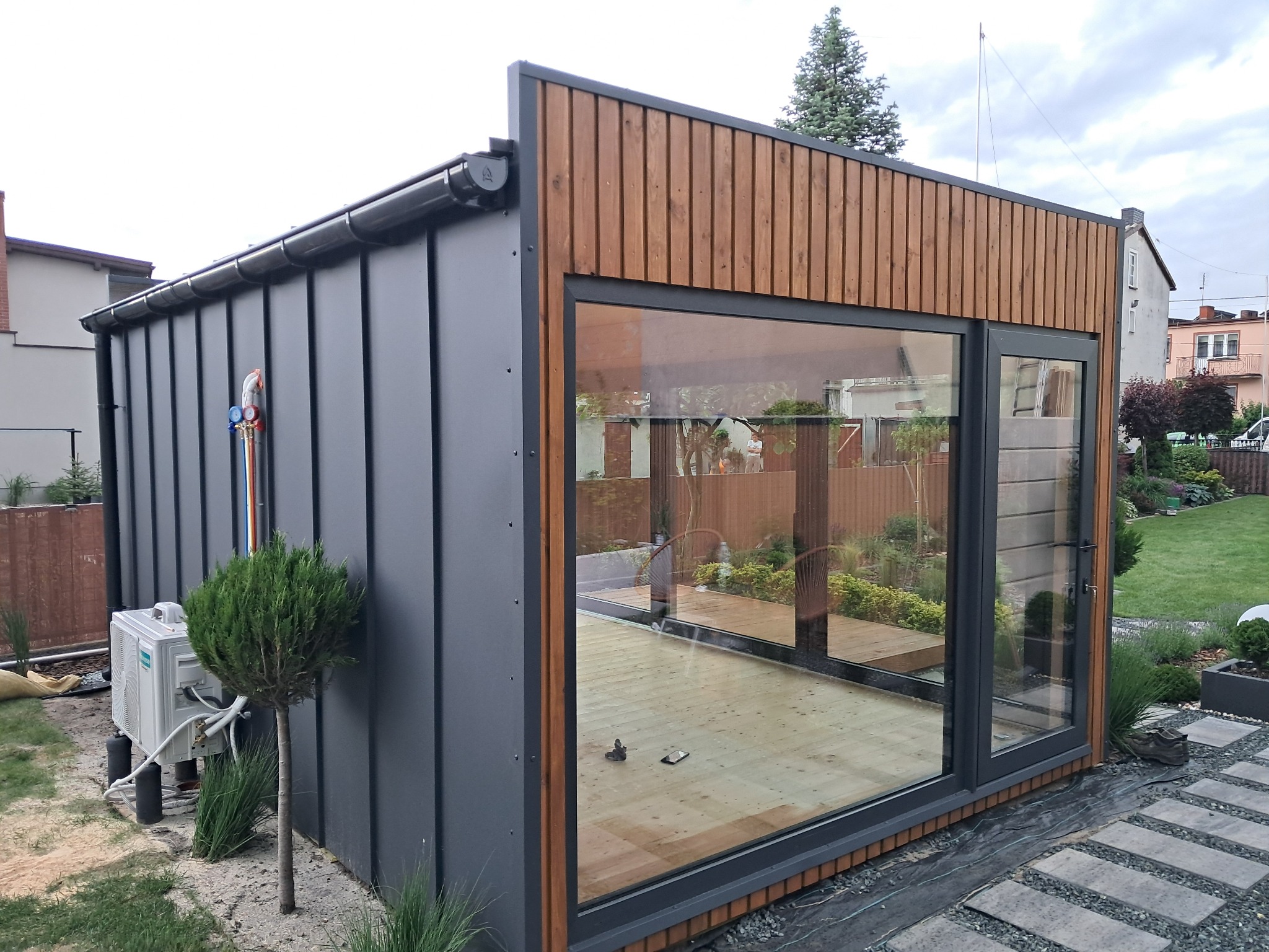 Nowoczesna, szara altana z drewnianymi elementami i dużym oknem, otoczona zielenią ogrodu. Widoczny klimatyzator na zewnątrz. Wnętrze altany z drewnianą podłogą.