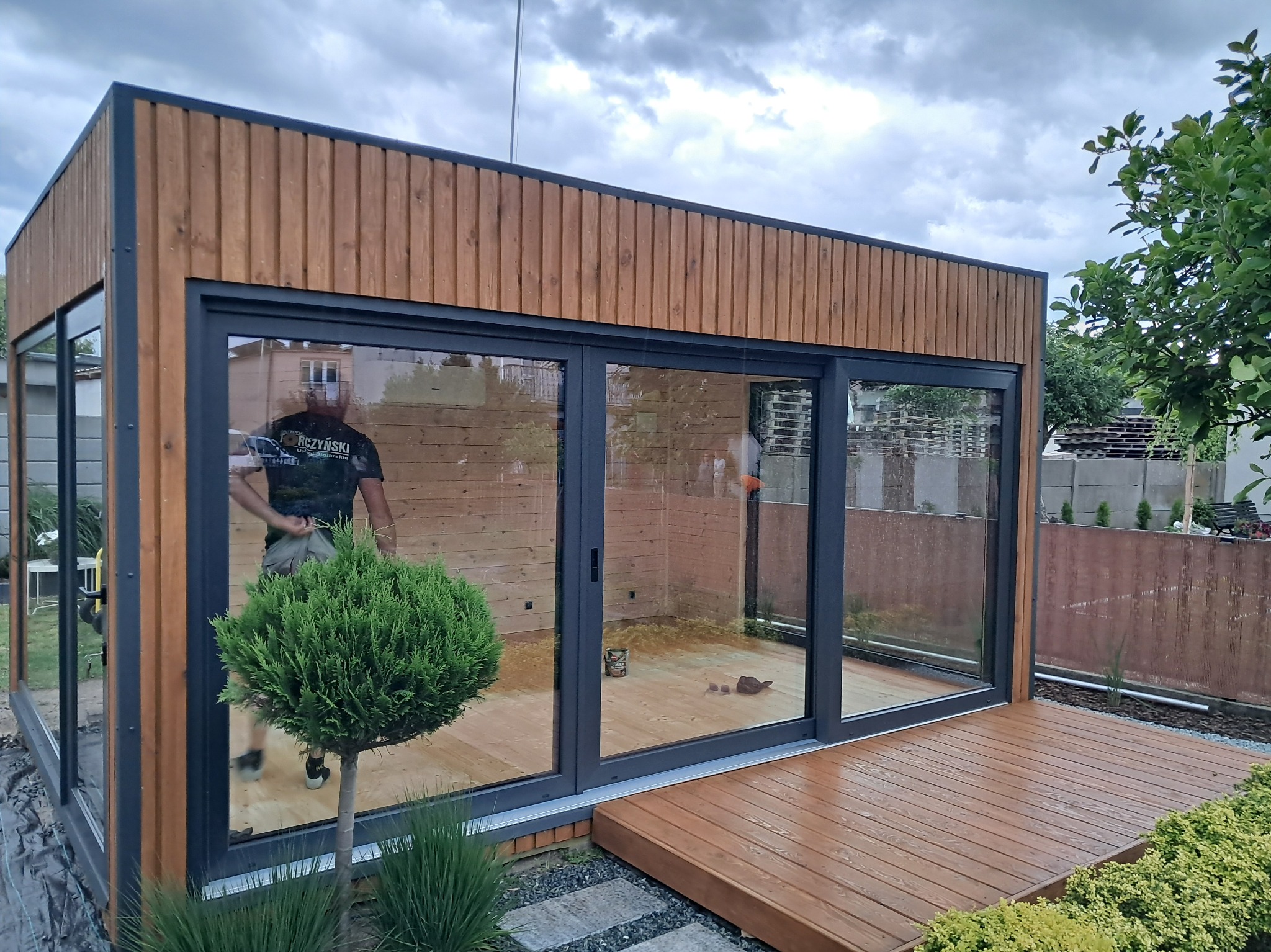 Nowoczesna, przeszklona altana z drewna, z przesuwnymi drzwiami i drewnianym tarasem. Widoczny fragment ogrodu i odbicie osoby w szybie.