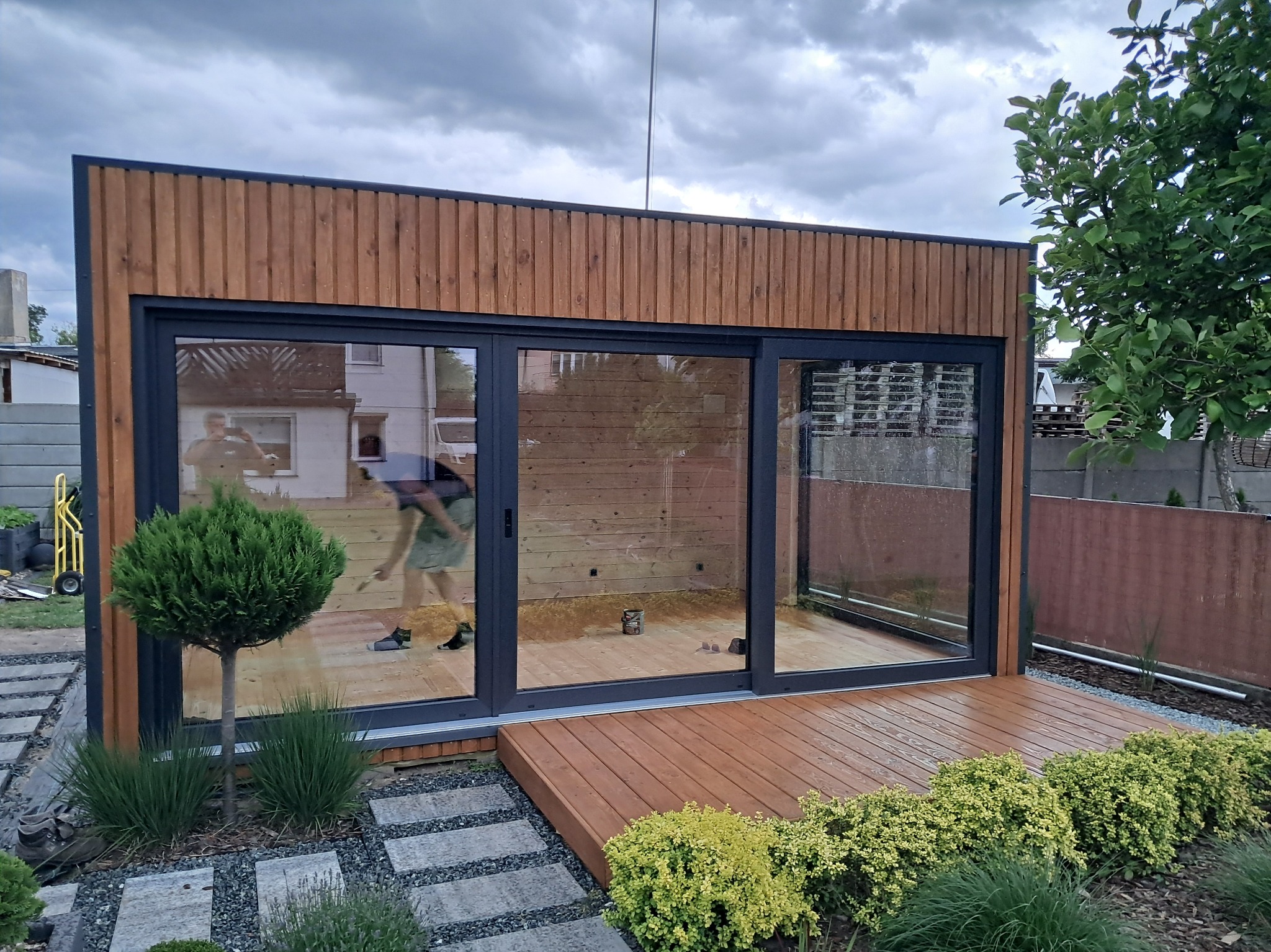 Nowoczesna altana z drewna i przeszkleniami, widoczna z zewnątrz. Taras z desek, rośliny ozdobne, ścieżka z kamieni. Prace wykończeniowe wewnątrz.