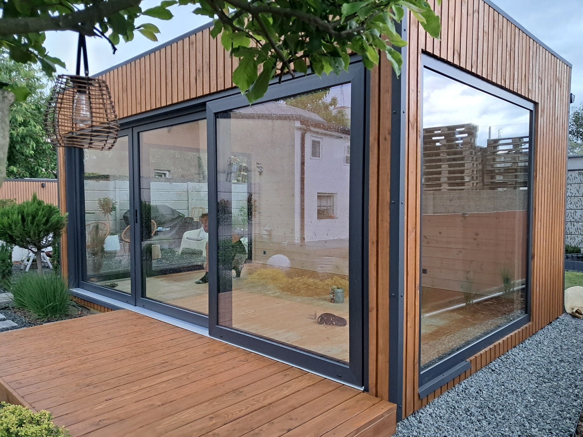 Nowoczesna altana z dużymi oknami przesuwnymi i drewnianym wykończeniem, zlokalizowana w Biskupicach, otoczona zielenią i żwirowym podłożem.