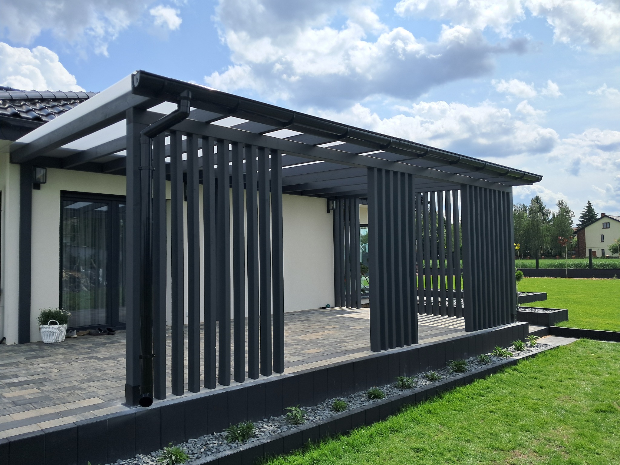 Nowoczesna pergola z grafitowymi, pionowymi lamelami i zadaszeniem, przylegająca do budynku. Widoczny taras z kostki i zadbany trawnik.