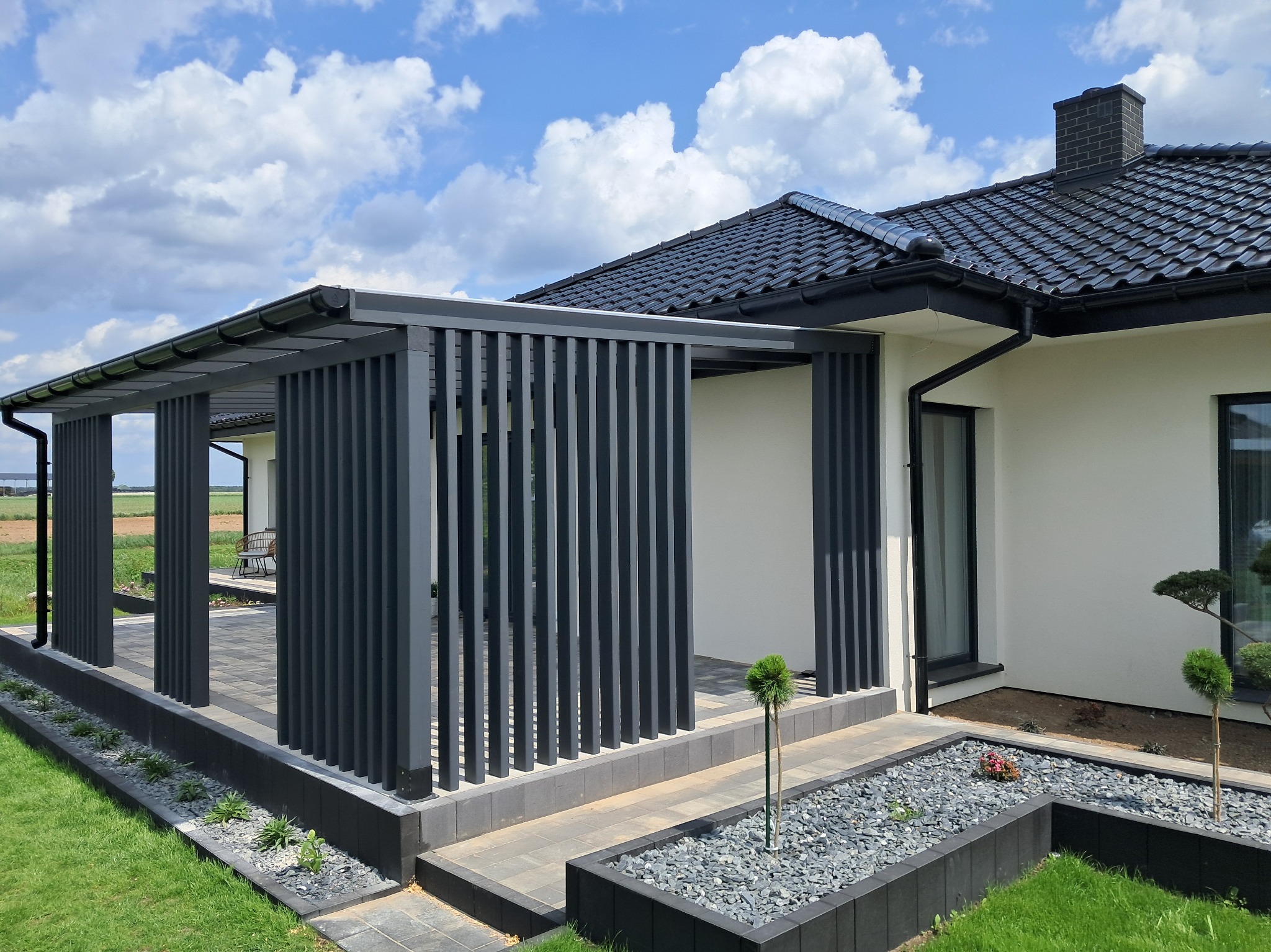 Nowoczesna, szara pergola z pionowymi lamelami przy białym domu z czarną dachówką. Widoczne zadbane trawniki i rabaty z kamieniami.