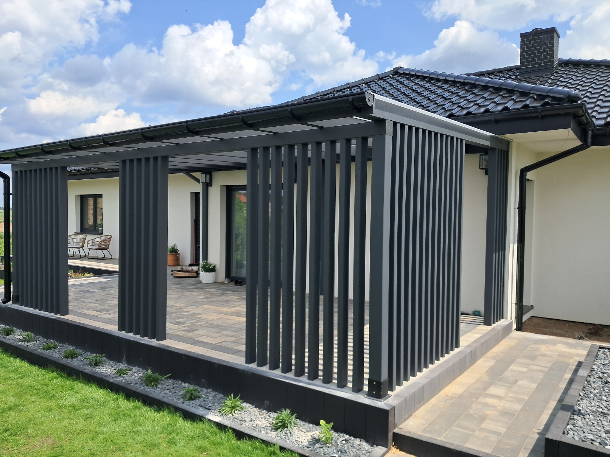 Nowoczesna pergola z szarymi, pionowymi panelami, zadaszeniem i kostką brukową. Element domu w tle. Detale wykończenia i aranżacji ogrodu.