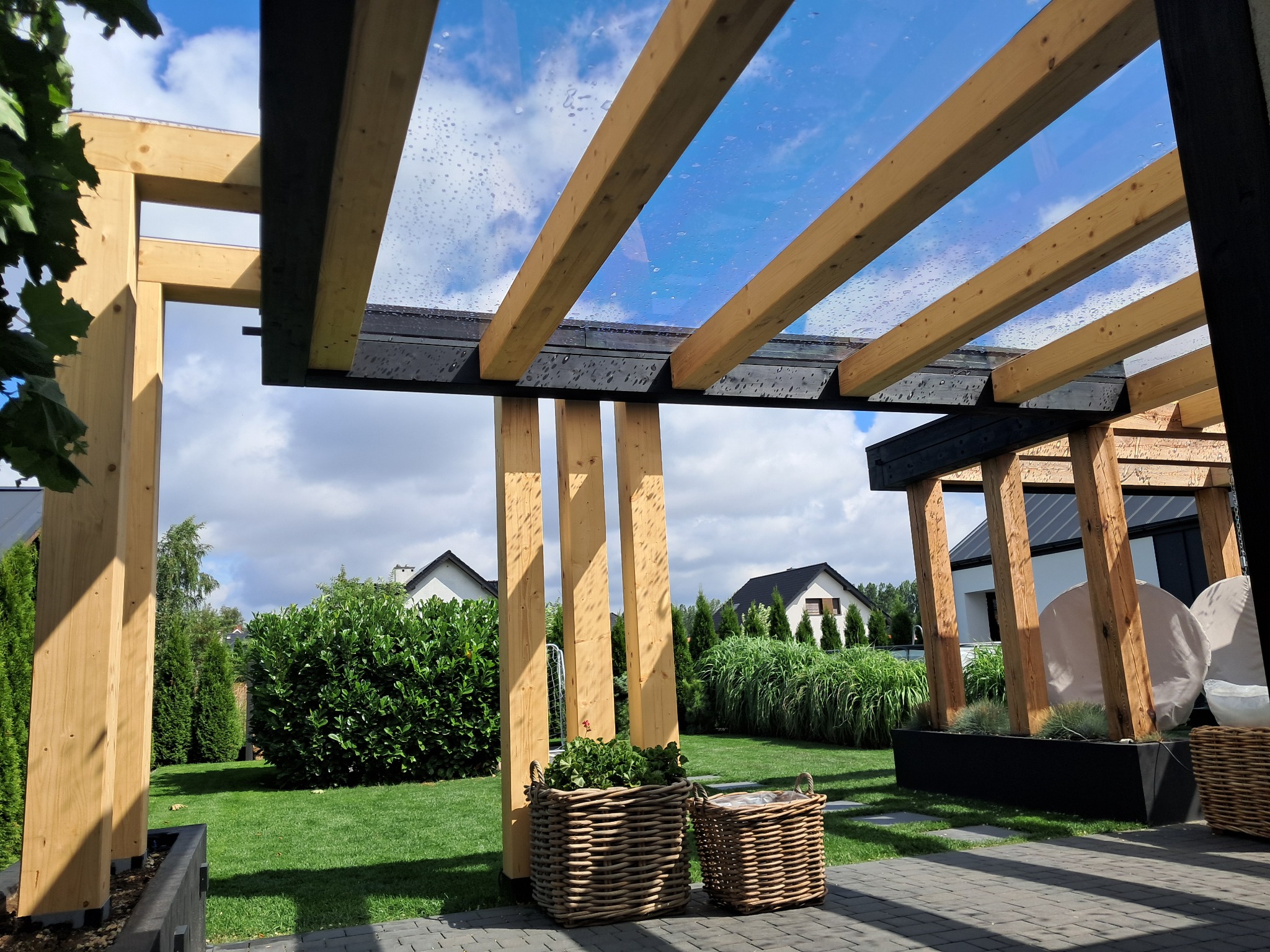 Nowoczesna pergola z drewna i szkła w ogrodzie, widok z dołu na konstrukcję i niebo. Dwa wiklinowe kosze z roślinami w tle, zadbany trawnik, domy w oddali.