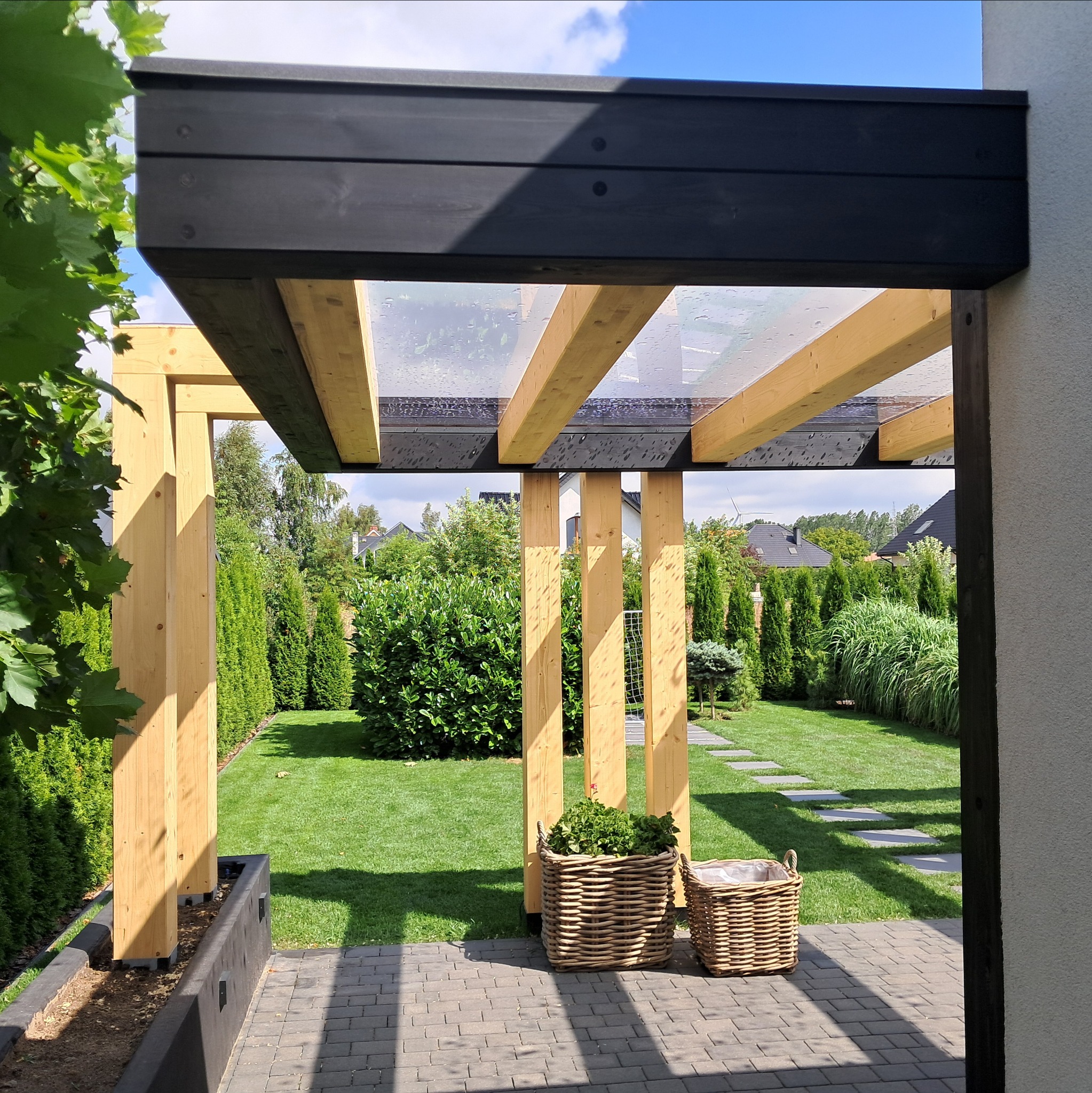 Drewniana pergola z przezroczystym zadaszeniem, widok na zadbany ogród z ścieżką. Dwa kosze wiklinowe z roślinami na pierwszym planie, nowoczesny design.