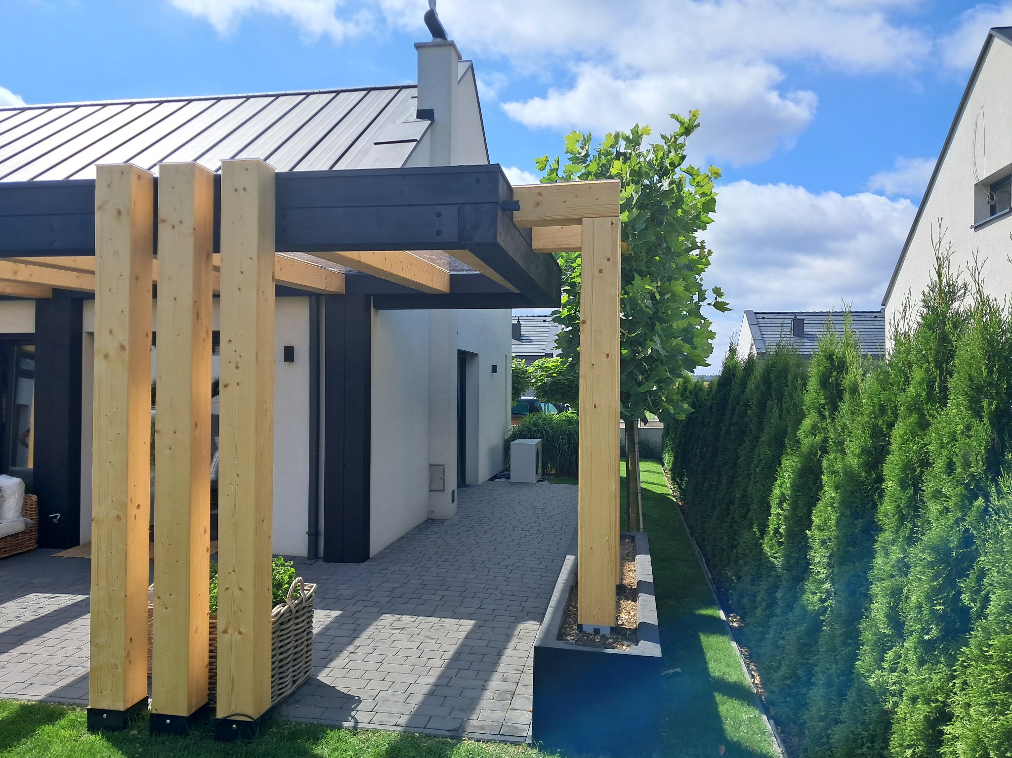 Nowoczesna pergola z jasnego drewna i czarnych belek na tle domu z szarą dachówką, otoczona zielenią. Widoczne detale konstrukcyjne i staranne wykonanie.