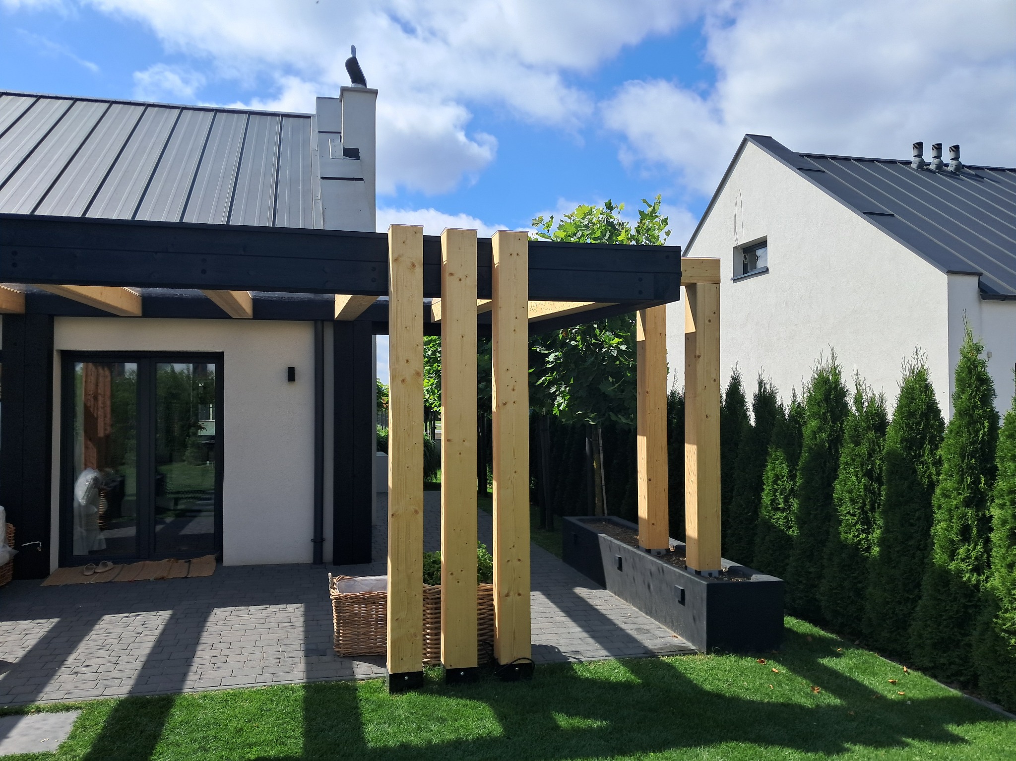 Nowoczesna pergola z jasnego drewna i czarnych belek na tle domu z szarą blachą trapezową, otoczona zielenią. Widoczny fragment tarasu z kostki.