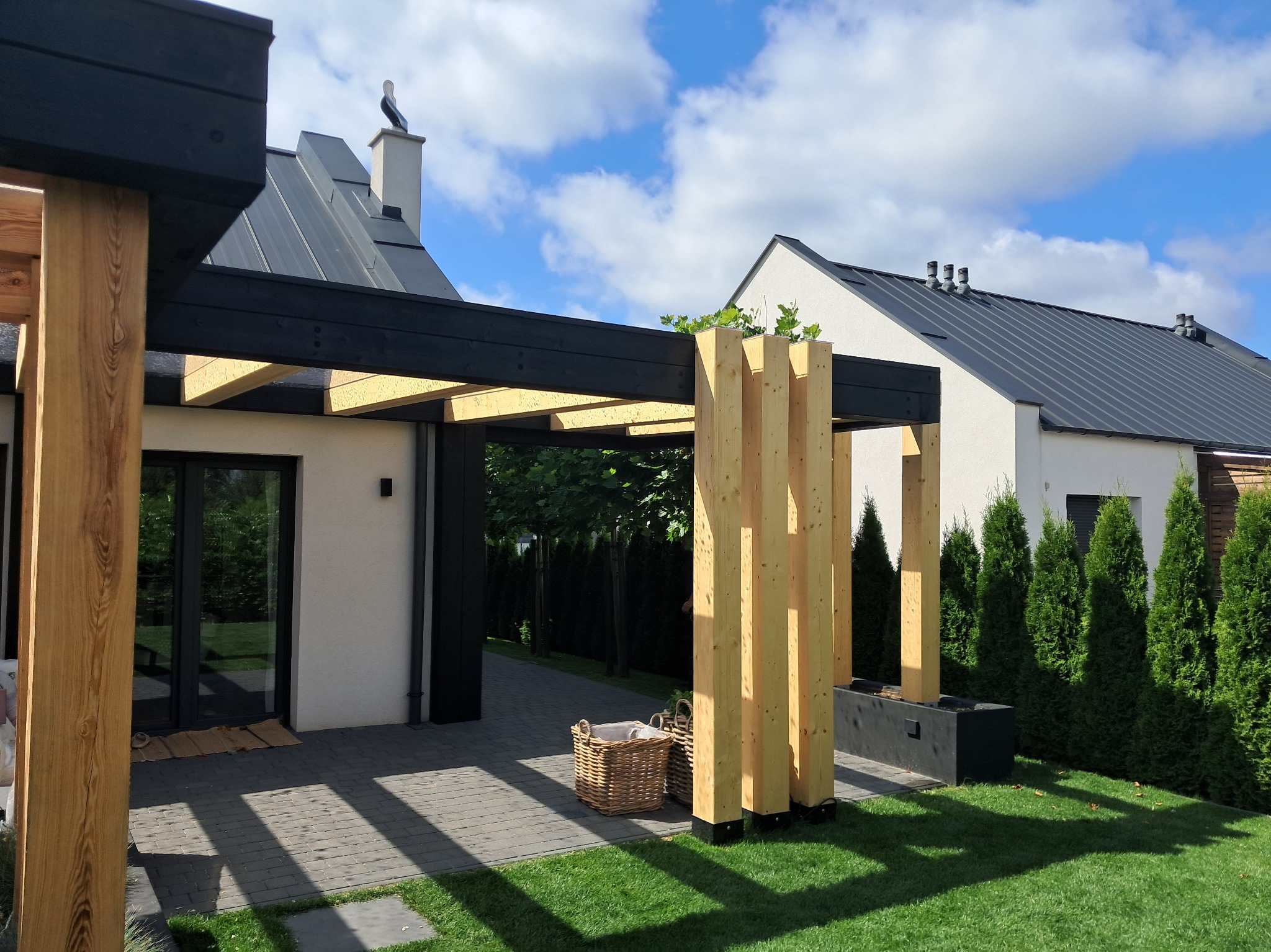 Nowoczesna pergola z jasnego drewna i czarnych belek na tle domu z szarą blachodachówką i zielonego trawnika. W pobliżu wiklinowy kosz i ozdobne tuje.