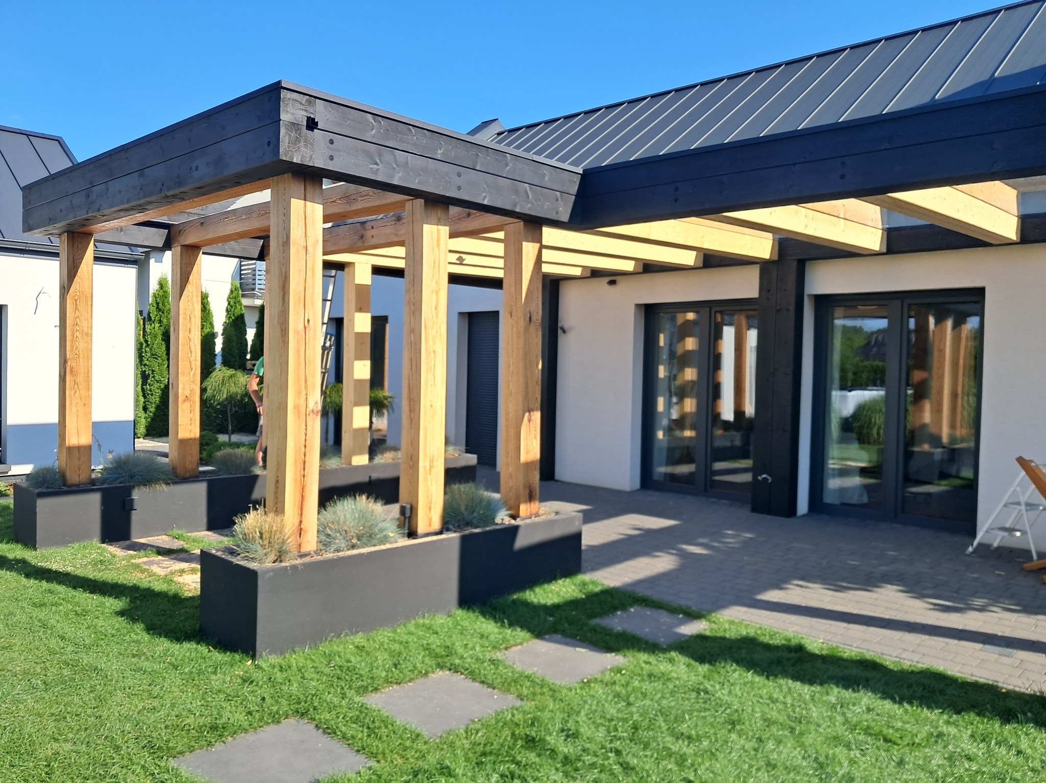 Nowoczesna pergola z drewnianymi słupami i czarną konstrukcją, zintegrowana z budynkiem. Minimalistyczne rabaty z roślinami ozdobnymi.