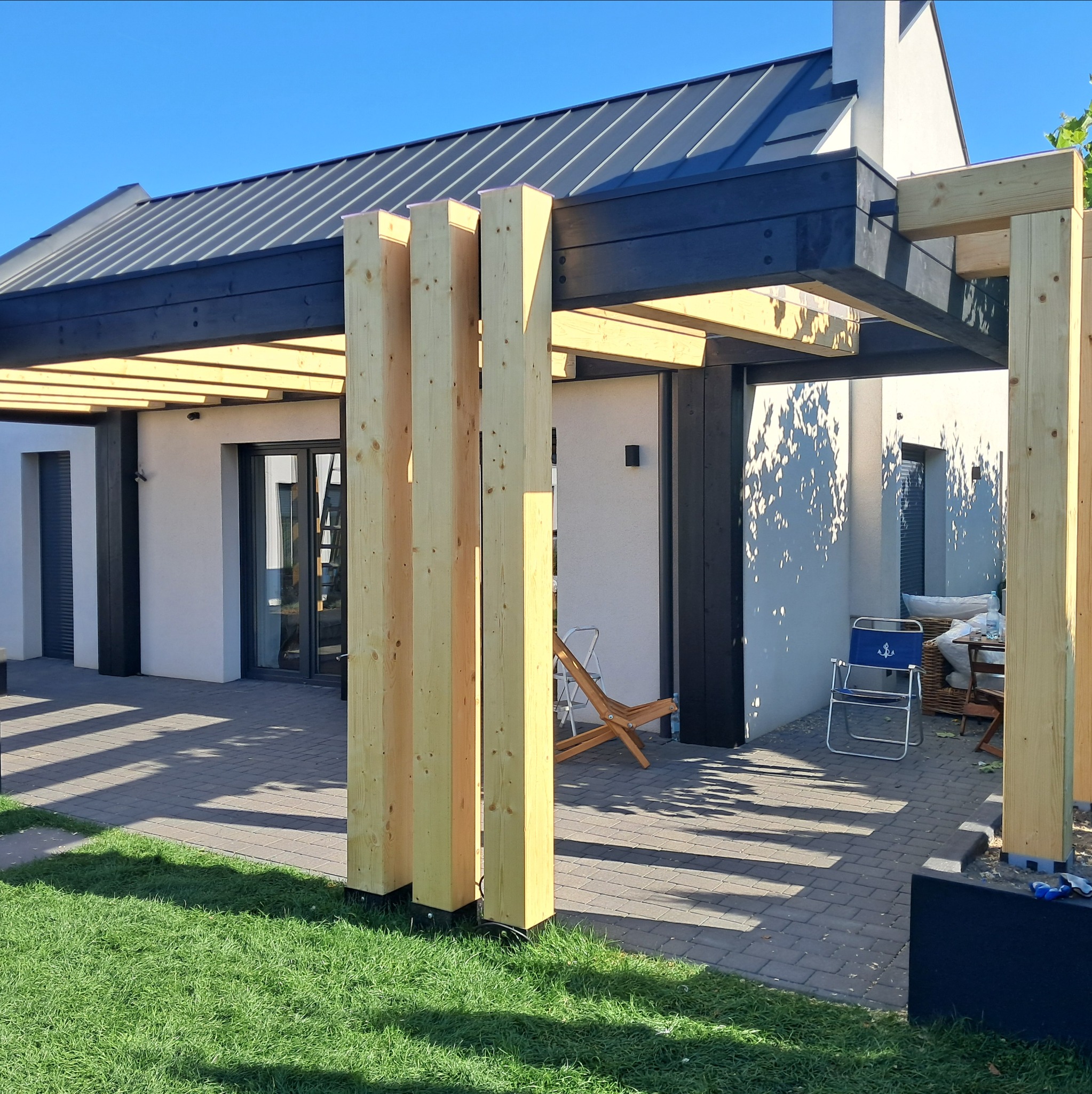 Nowoczesna pergola z jasnego drewna i ciemnych belek na tle domu z szarą blachodachówką. Widoczne meble ogrodowe i trawnik.