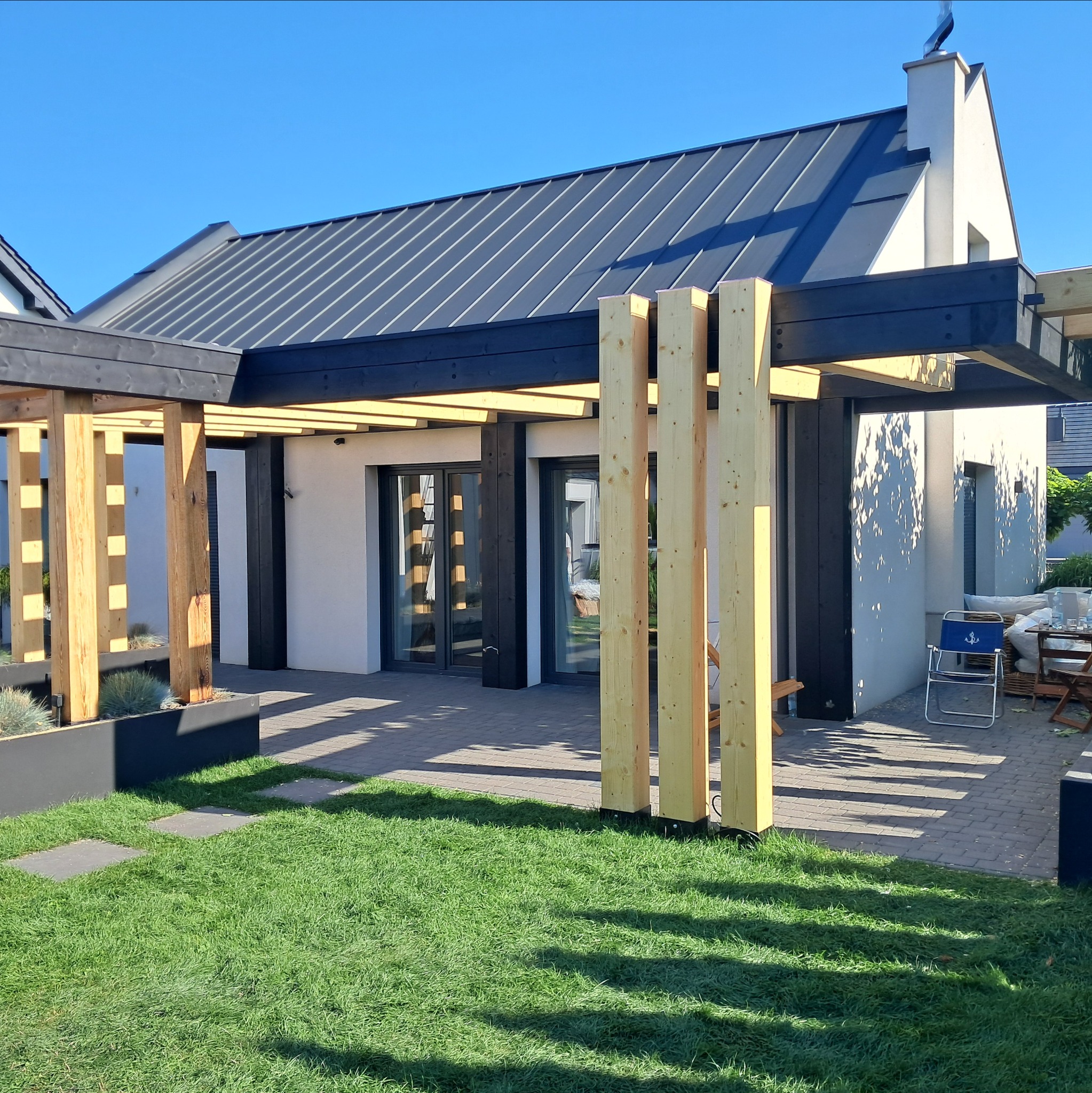Nowoczesna pergola z surowego drewna i czarnych belek przy białym domu z szarym dachem, widok z trawnika. Elementy architektury ogrodowej w słoneczny dzień.