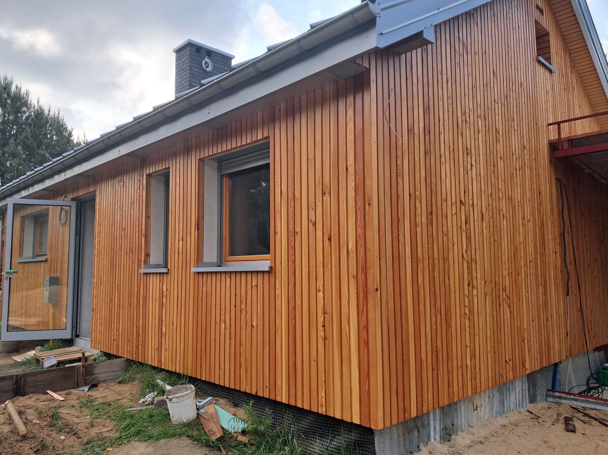 Nowoczesny dom szkieletowy z elewacją z pionowo ułożonych desek w ciepłym, miodowym odcieniu drewna. Widoczne okna i otwarte drzwi wejściowe. W tle komin i fragment rusztowania.