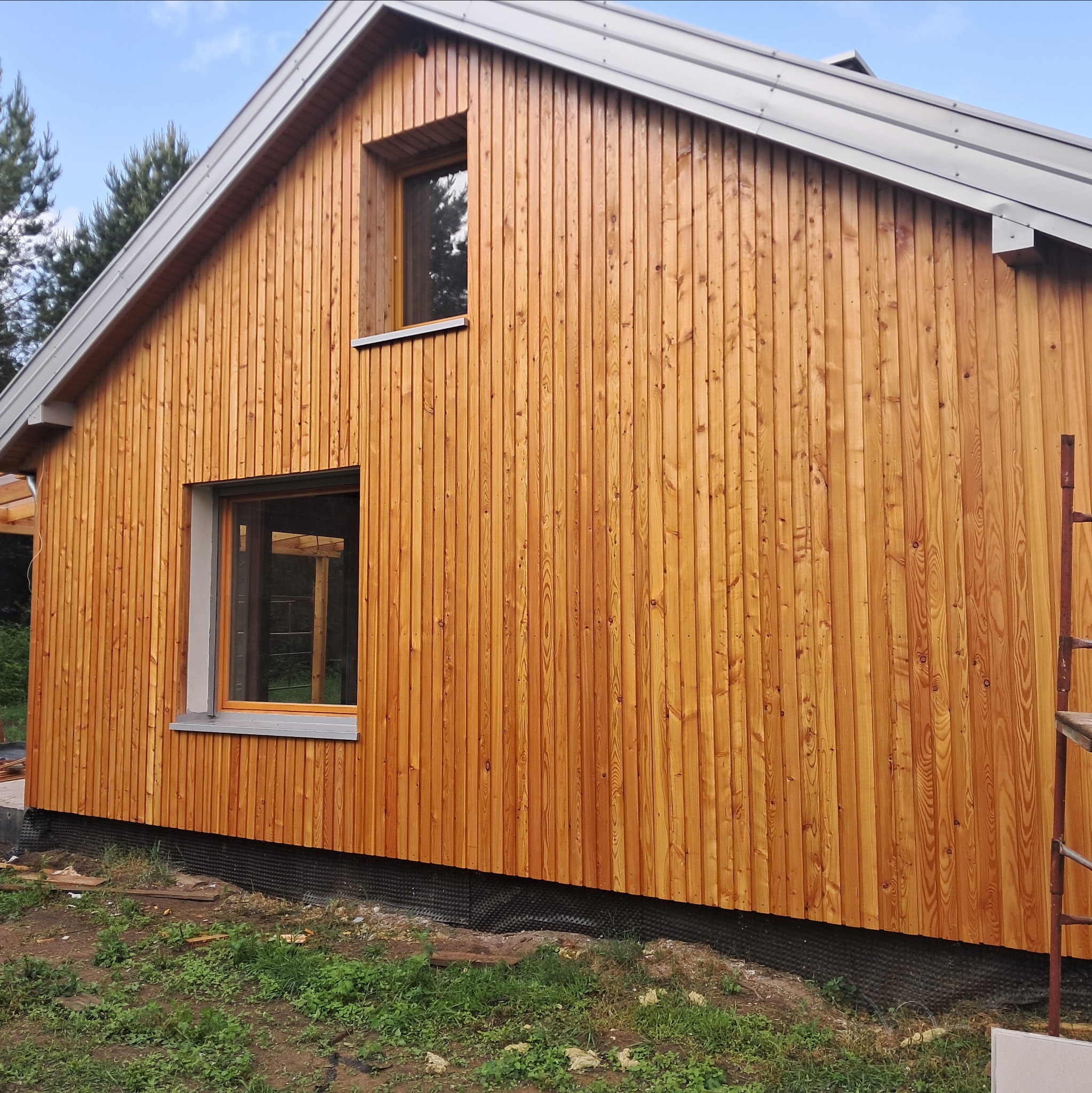 Dom szkieletowy z jasną, drewnianą elewacją pionową i szarym dachem. Widoczne dwa okna i fragment rusztowania. W tle drzewa i błękitne niebo.