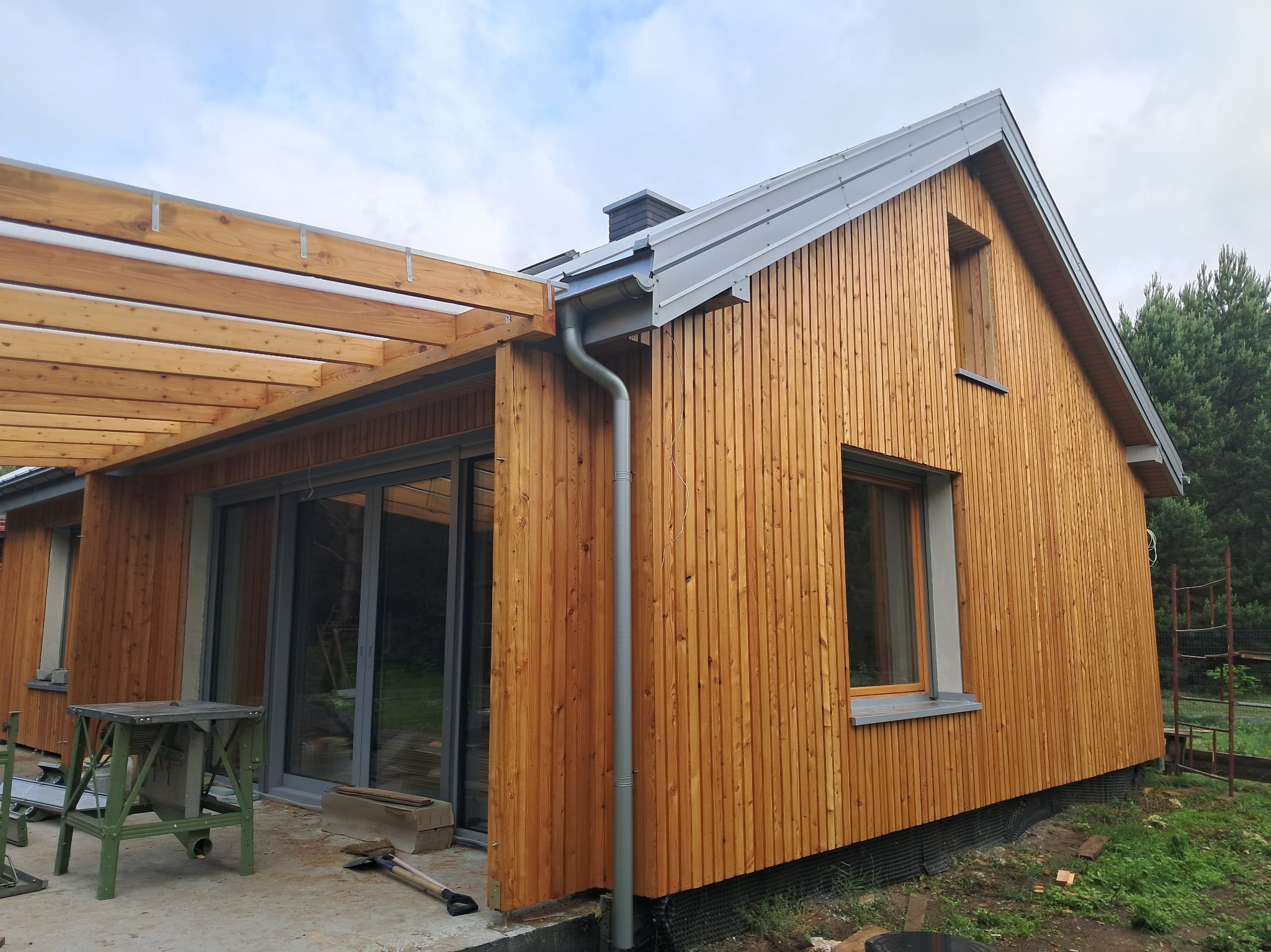 Nowoczesny dom szkieletowy z drewnianą elewacją i pergolą, widok z zewnątrz. Detale: rynna, okna, narzędzia na placu budowy. Jasne drewno, metalowy dach.