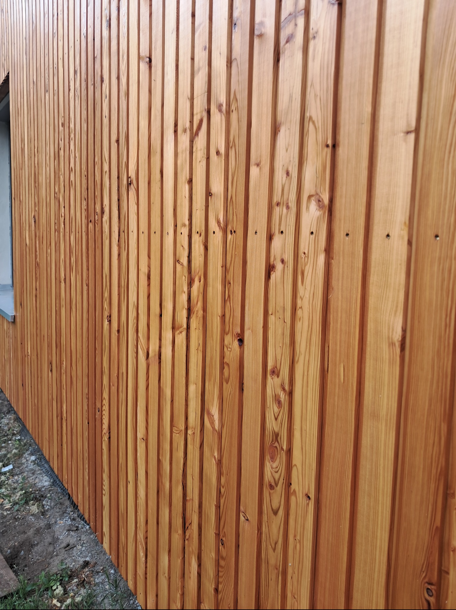 Elewacja budynku wykończona pionowymi deskami z jasnego drewna, ukazująca naturalny rysunek słojów. Widoczny fragment okna z parapetem i przygruntowy teren.