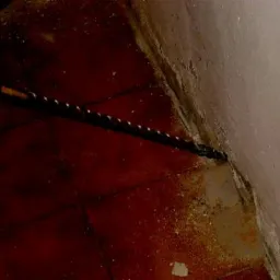 Wiertło w ścianie, widoczne zawilgocenie i uszkodzenia na styku ściany z podłogą z czerwonej terakoty.