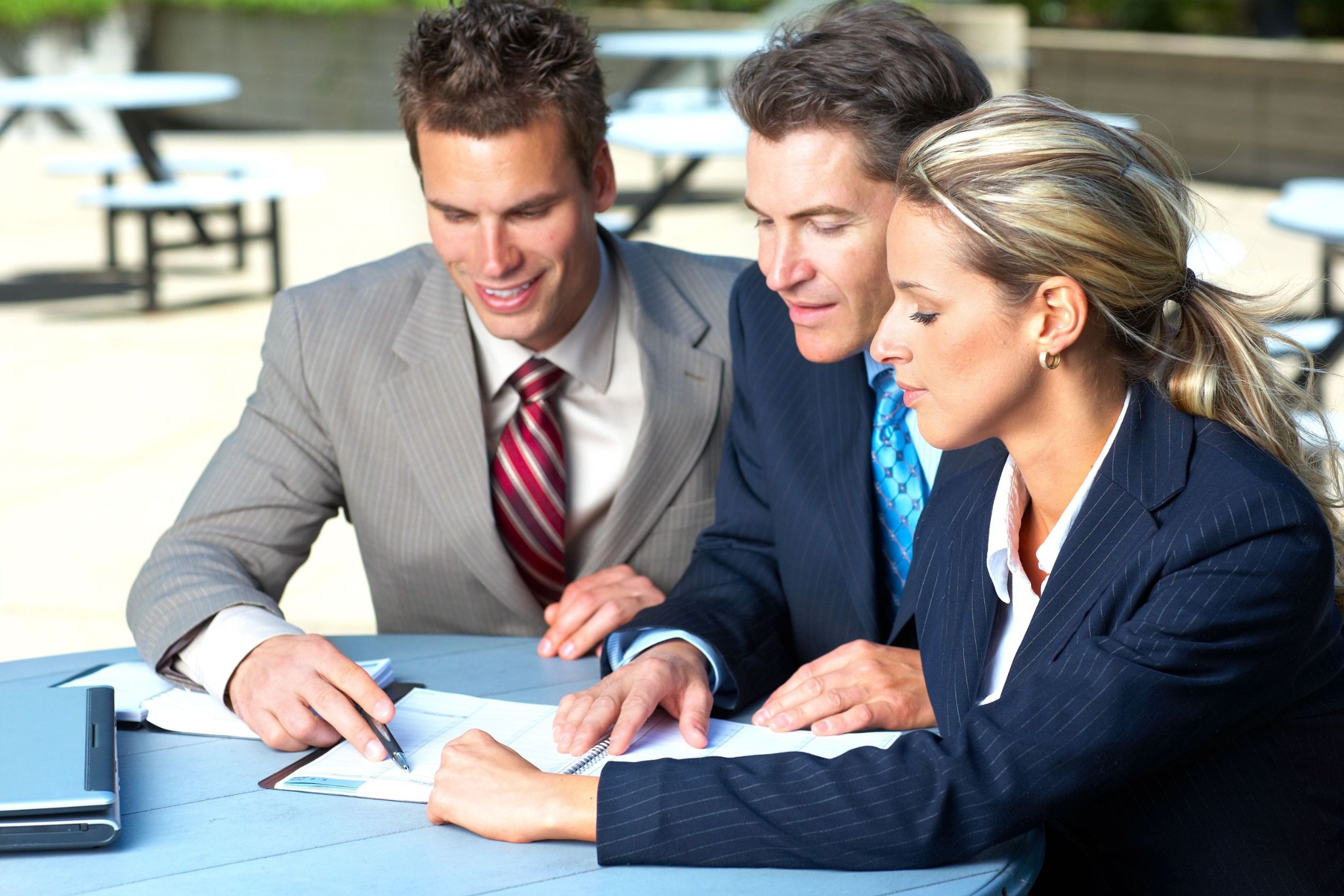 Trzech profesjonalistów w garniturach pochyla się nad dokumentami na stole, jeden z nich trzyma długopis, obok leży laptop, w tle stoliki na zewnątrz.