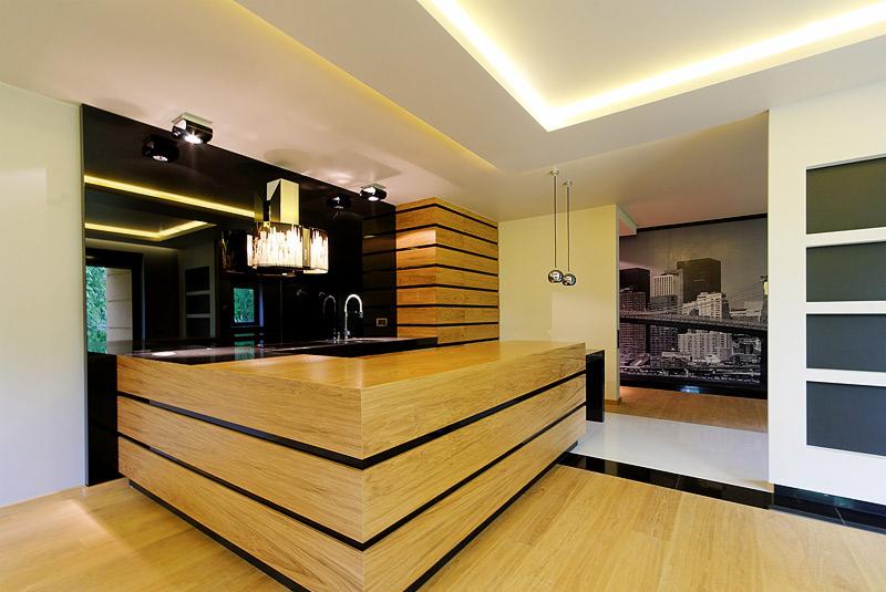 Nowoczesna kuchnia z wyspą z jasnego drewna, czarnym blatem i lustrzanym panelem ściennym, oświetlenie LED podsufitowe, w tle widoczna fototapeta z motywem miejskim.